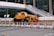A yellow crane truck is parked in an urban construction site, surrounded by orange and white traffic cones and barricades. A pedestrian bridge arches overhead, and a modern building façade is visible in the background.