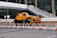 A yellow crane truck is parked in an urban construction site, surrounded by orange and white traffic cones and barricades. A pedestrian bridge arches overhead, and a modern building façade is visible in the background.