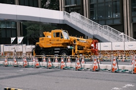 A yellow crane truck is parked in an urban construction site, surrounded by orange and white traffic cones and barricades. A pedestrian bridge arches overhead, and a modern building façade is visible in the background.