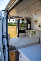 The interior of a camper van is depicted with a cozy setup including a bed, soft pillows, and a hanging string of small lights. The bed is adorned with a pillow featuring an image of a butterfly. The van door is open, giving a view of a grassy field and trees outside. The interior appears clean and minimalistic, with neutral tones and a small cabinet in view.