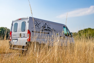 A white van is parked in a field of tall, dry grass under a clear blue sky. The van has a large graphic of a compass and mountain range on its side. Surrounding the field are trees in the distance, suggesting a rural or remote location.