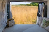 A cozy interior of a van with a bed facing an open door, revealing a serene view of a grassy field. A dreamcatcher and pillows with nature-themed designs add to the tranquil atmosphere. The horizon displays a line of trees and a clear sky.
