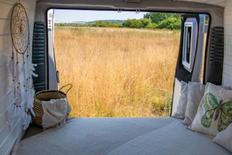A cozy interior of a van with a bed facing an open door, revealing a serene view of a grassy field. A dreamcatcher and pillows with nature-themed designs add to the tranquil atmosphere. The horizon displays a line of trees and a clear sky.