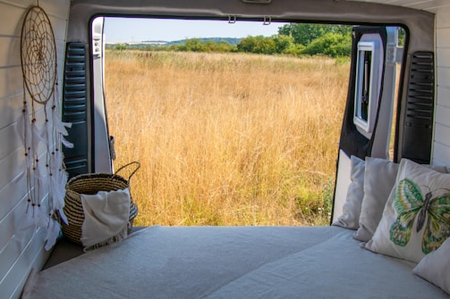 A cozy interior of a van with a bed facing an open door, revealing a serene view of a grassy field. A dreamcatcher and pillows with nature-themed designs add to the tranquil atmosphere. The horizon displays a line of trees and a clear sky.