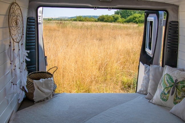 A cozy interior of a van with a bed facing an open door, revealing a serene view of a grassy field. A dreamcatcher and pillows with nature-themed designs add to the tranquil atmosphere. The horizon displays a line of trees and a clear sky.