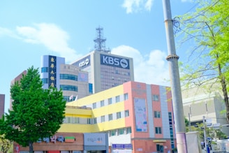 A modern urban scene with several multi-story buildings featuring a mix of architectural styles. Prominent signage with the text 'KBS' indicates a media or broadcasting company. The foreground includes a colorful building with yellow and pink hues, and commercial establishments at the street level. Various trees with lush green foliage frame the scene, with a clear blue sky overhead.