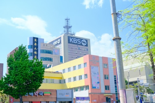 A modern urban scene with several multi-story buildings featuring a mix of architectural styles. Prominent signage with the text 'KBS' indicates a media or broadcasting company. The foreground includes a colorful building with yellow and pink hues, and commercial establishments at the street level. Various trees with lush green foliage frame the scene, with a clear blue sky overhead.