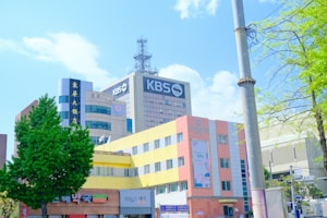 A modern urban scene with several multi-story buildings featuring a mix of architectural styles. Prominent signage with the text 'KBS' indicates a media or broadcasting company. The foreground includes a colorful building with yellow and pink hues, and commercial establishments at the street level. Various trees with lush green foliage frame the scene, with a clear blue sky overhead.