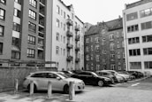 A collection of parked vehicles is located in front of several tall, multi-story apartment buildings, with some structures featuring brick facades and others having plain plaster exteriors. The image appears to capture an urban residential area with a mix of modern and older architectural elements.