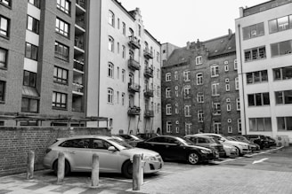 A collection of parked vehicles is located in front of several tall, multi-story apartment buildings, with some structures featuring brick facades and others having plain plaster exteriors. The image appears to capture an urban residential area with a mix of modern and older architectural elements.
