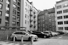 A collection of parked vehicles is located in front of several tall, multi-story apartment buildings, with some structures featuring brick facades and others having plain plaster exteriors. The image appears to capture an urban residential area with a mix of modern and older architectural elements.
