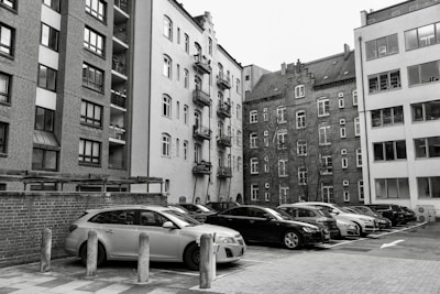 A collection of parked vehicles is located in front of several tall, multi-story apartment buildings, with some structures featuring brick facades and others having plain plaster exteriors. The image appears to capture an urban residential area with a mix of modern and older architectural elements.
