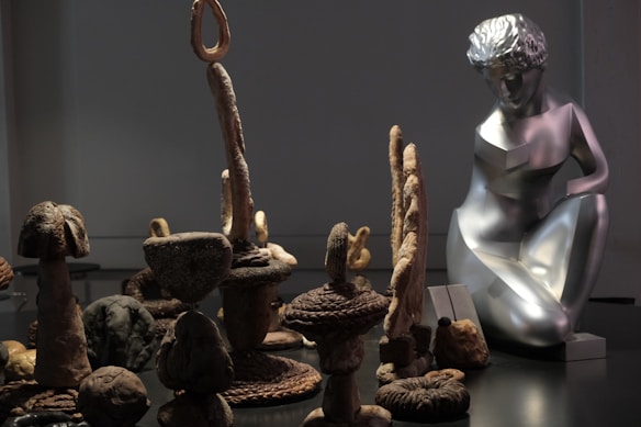 A collection of abstract sculptures and art pieces arranged in a dimly lit space. On the right, a metallic sculpture of a human figure is prominently displayed, characterized by stylized features and a reflective surface. Surrounding it are various organic-shaped structures made of materials resembling stone and wood, with intricate textures and natural forms.