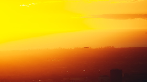 A sleek modern airplane flying over a vibrant city skyline at sunset.