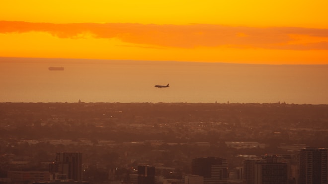A cityscape at sunset featuring a silhouette of an airplane flying above the urban skyline. The sky is a warm gradient of orange and yellow hues, with a distant ship visible on the horizon.