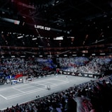 A large indoor stadium filled with spectators, with a central tennis court in view. Two players are on the court, and the court is surrounded by LED displays bearing the "Laver Cup" branding. The audience is vibrant, with sections lit in blue and red, creating a dramatic atmosphere.