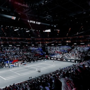 A large indoor stadium filled with spectators, with a central tennis court in view. Two players are on the court, and the court is surrounded by LED displays bearing the "Laver Cup" branding. The audience is vibrant, with sections lit in blue and red, creating a dramatic atmosphere.