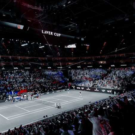 A large indoor stadium filled with spectators, with a central tennis court in view. Two players are on the court, and the court is surrounded by LED displays bearing the "Laver Cup" branding. The audience is vibrant, with sections lit in blue and red, creating a dramatic atmosphere.