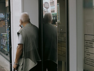 An elderly person with white hair, wearing a mask, a gray short-sleeved shirt, and a vest, is standing outside a store or office building, near a glass door. They are partially reflected in the glass, and they're holding a small object in one hand. In the background, a bus or large vehicle is slightly visible through the reflection on the door.