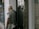 An elderly person with white hair, wearing a mask, a gray short-sleeved shirt, and a vest, is standing outside a store or office building, near a glass door. They are partially reflected in the glass, and they're holding a small object in one hand. In the background, a bus or large vehicle is slightly visible through the reflection on the door.