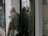 An elderly person with white hair, wearing a mask, a gray short-sleeved shirt, and a vest, is standing outside a store or office building, near a glass door. They are partially reflected in the glass, and they're holding a small object in one hand. In the background, a bus or large vehicle is slightly visible through the reflection on the door.
