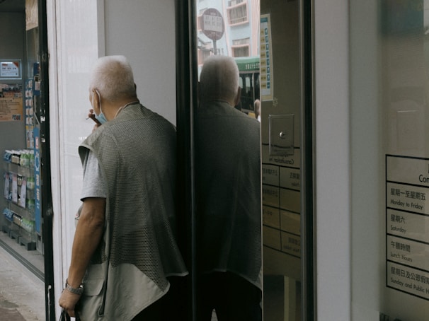 An elderly person with white hair, wearing a mask, a gray short-sleeved shirt, and a vest, is standing outside a store or office building, near a glass door. They are partially reflected in the glass, and they're holding a small object in one hand. In the background, a bus or large vehicle is slightly visible through the reflection on the door.