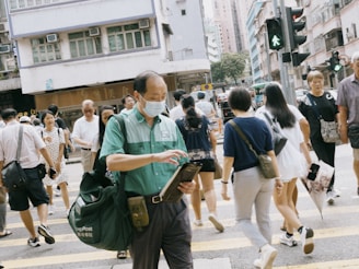 A bustling urban scene with people crossing the street at a pedestrian crossing in a city. Most are casually dressed, and at the forefront, a person wearing a green uniform and face mask appears to be checking a clipboard. The background shows buildings, traffic lights, and signs.