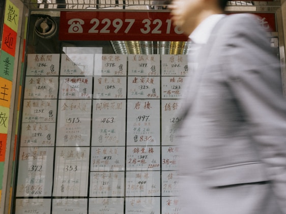 A person in a gray suit is blurred as they walk past a window displaying numerous pages with Chinese characters and numbers, possibly indicating advertisements or listings. The top of the window displays a red banner with a phone number in white. Brightly colored paper strips with characters are visible on the left side of the frame.