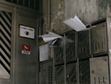 A group of metallic mailboxes attached to a worn, dirty wall. Some mailboxes have envelopes and papers sticking out. Nearby, there is a door with various signs in another language including an eye symbol and a red sign indicating 'No Smoking'.