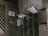 A group of metallic mailboxes attached to a worn, dirty wall. Some mailboxes have envelopes and papers sticking out. Nearby, there is a door with various signs in another language including an eye symbol and a red sign indicating 'No Smoking'.