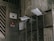A group of metallic mailboxes attached to a worn, dirty wall. Some mailboxes have envelopes and papers sticking out. Nearby, there is a door with various signs in another language including an eye symbol and a red sign indicating 'No Smoking'.