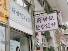 A street view of a building facade displays a barber's shop sign in Chinese characters. The sign is prominently placed above the entrance along with a traditional barber pole featuring red, white, and blue stripes. Nearby, another sign offers haircut prices, and the storefront has a window with promotional images.