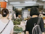 A small local market stall with a vendor and two customers. The stall is filled with various vegetables and herbs, and there are signs with prices written in a foreign language. The vendor appears to be an elderly person, situated in a narrow space surrounded by goods. The two customers are facing the stall, possibly deciding on their purchase.