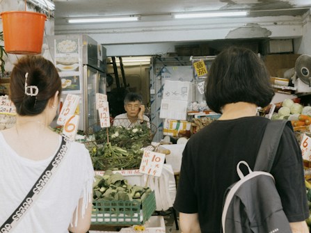 A small local market stall with a vendor and two customers. The stall is filled with various vegetables and herbs, and there are signs with prices written in a foreign language. The vendor appears to be an elderly person, situated in a narrow space surrounded by goods. The two customers are facing the stall, possibly deciding on their purchase.