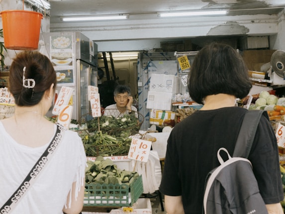 A small local market stall with a vendor and two customers. The stall is filled with various vegetables and herbs, and there are signs with prices written in a foreign language. The vendor appears to be an elderly person, situated in a narrow space surrounded by goods. The two customers are facing the stall, possibly deciding on their purchase.