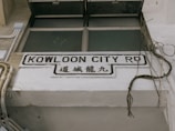 A street sign with the text 'Kowloon City Rd' in English and corresponding Chinese characters below it, mounted on a white concrete wall. Surrounding the sign are some exposed electrical wires and pipes, suggesting an urban setting.