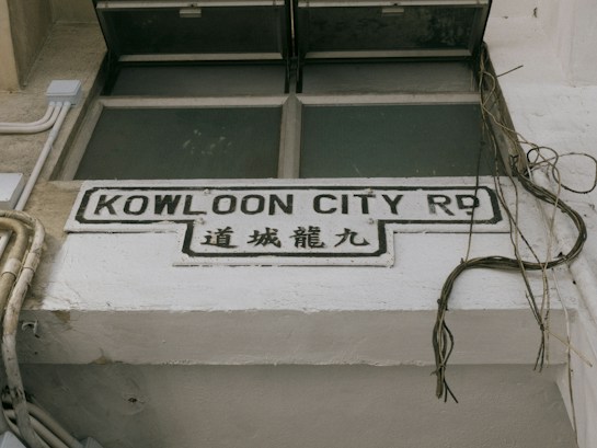 A street sign with the text 'Kowloon City Rd' in English and corresponding Chinese characters below it, mounted on a white concrete wall. Surrounding the sign are some exposed electrical wires and pipes, suggesting an urban setting.