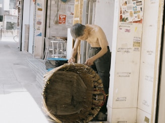 An elderly man stands in a narrow alleyway, handling a large woven basket. He is shirtless and seems focused on the task. The surroundings include a tiled floor, walls with posters, and various objects like chairs and containers. The area appears to be part of a residential or market setting.