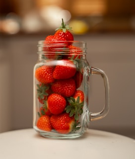 Close-up of a jar of Maumeladas strawberry jam with fresh strawberries around it on a rustic wooden table.