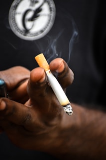 A hand holds a burning cigarette with smoke rising from the tip. The background is dark, and there is a circular logo visible, slightly blurred.