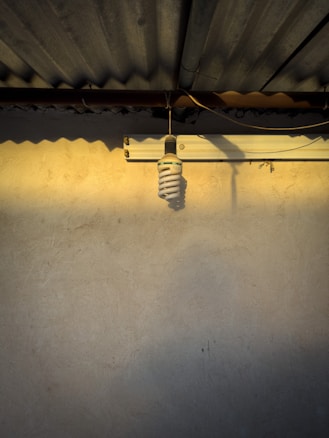 A spiral-shaped compact fluorescent light bulb hangs from the ceiling. The ceiling has corrugated metal sheets, casting shadows on a textured beige wall. Warm sunlight creates a gradient of light across the surface.