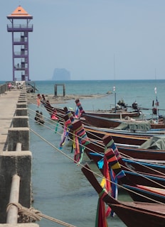 A series of traditional wooden boats are tied to a pier, adorned with colorful ribbons. The ocean stretches into the background, with a tall purple observation tower adjacent to the pier. In the distance, a small island or rock formation appears on the horizon under a clear blue sky.