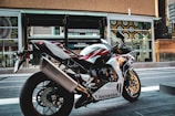 A sleek Honda motorcycle is parked on an urban street. The design of the motorcycle features a sporty build with a color scheme of white, red, and blue. It is positioned in front of a modern building with large windows and decorative patterns.