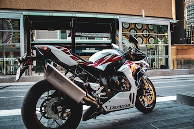 A sleek Honda motorcycle is parked on an urban street. The design of the motorcycle features a sporty build with a color scheme of white, red, and blue. It is positioned in front of a modern building with large windows and decorative patterns.
