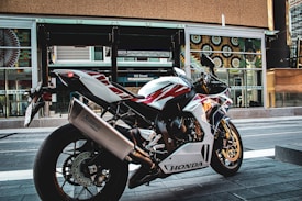 A sleek Honda motorcycle is parked on an urban street. The design of the motorcycle features a sporty build with a color scheme of white, red, and blue. It is positioned in front of a modern building with large windows and decorative patterns.