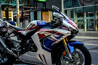 A detailed and colorful Honda motorcycle is parked on a city street. The motorcycle is adorned with a combination of white, blue, red, and black colors. It has a sleek design, with visible branding and graphics on the body. The background shows a storefront with reflective windows, a street sign, and some urban elements such as pedestrian paths and a bus stop.