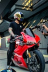 A person wearing a helmet with intricate designs sits on a bright red Ducati motorcycle in a modern urban setting. The individual is dressed in a black t-shirt and has tattoos on their arms. Another person, partially visible, appears to be filming with a camera mounted on a stabilizer.