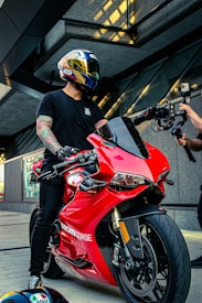 A person wearing a helmet with intricate designs sits on a bright red Ducati motorcycle in a modern urban setting. The individual is dressed in a black t-shirt and has tattoos on their arms. Another person, partially visible, appears to be filming with a camera mounted on a stabilizer.