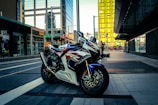 A motorcycle with a sporty design parked on a modern city street, surrounded by tall buildings with reflective glass surfaces. The environment shows a blend of urban architecture and outdoor elements, with a person in the background near a storefront.