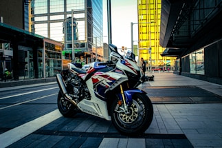 A motorcycle with a sporty design parked on a modern city street, surrounded by tall buildings with reflective glass surfaces. The environment shows a blend of urban architecture and outdoor elements, with a person in the background near a storefront.
